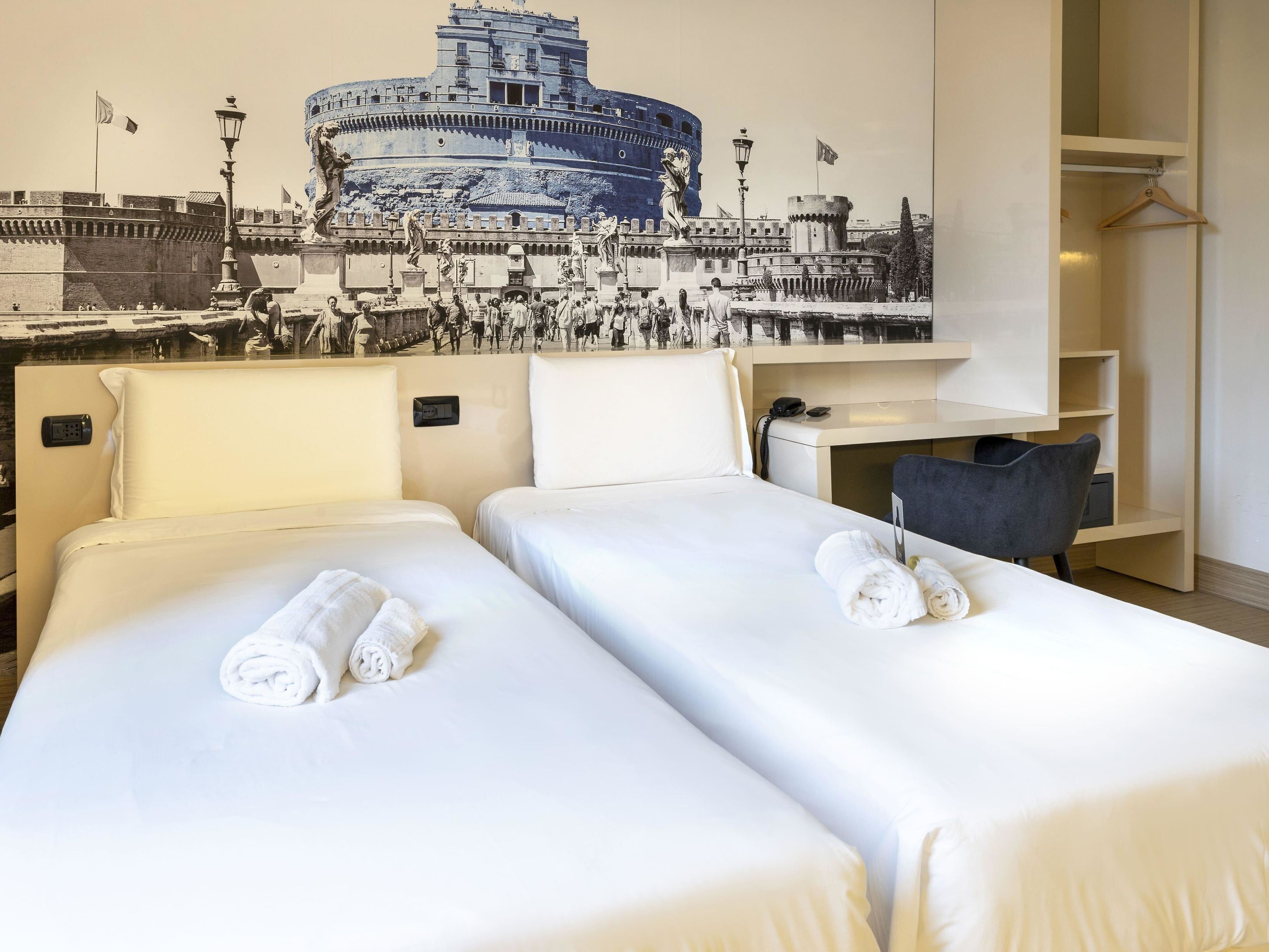 Habitación de hotel con dos camas, un escritorio, una silla y un mural grande de un edificio histórico.