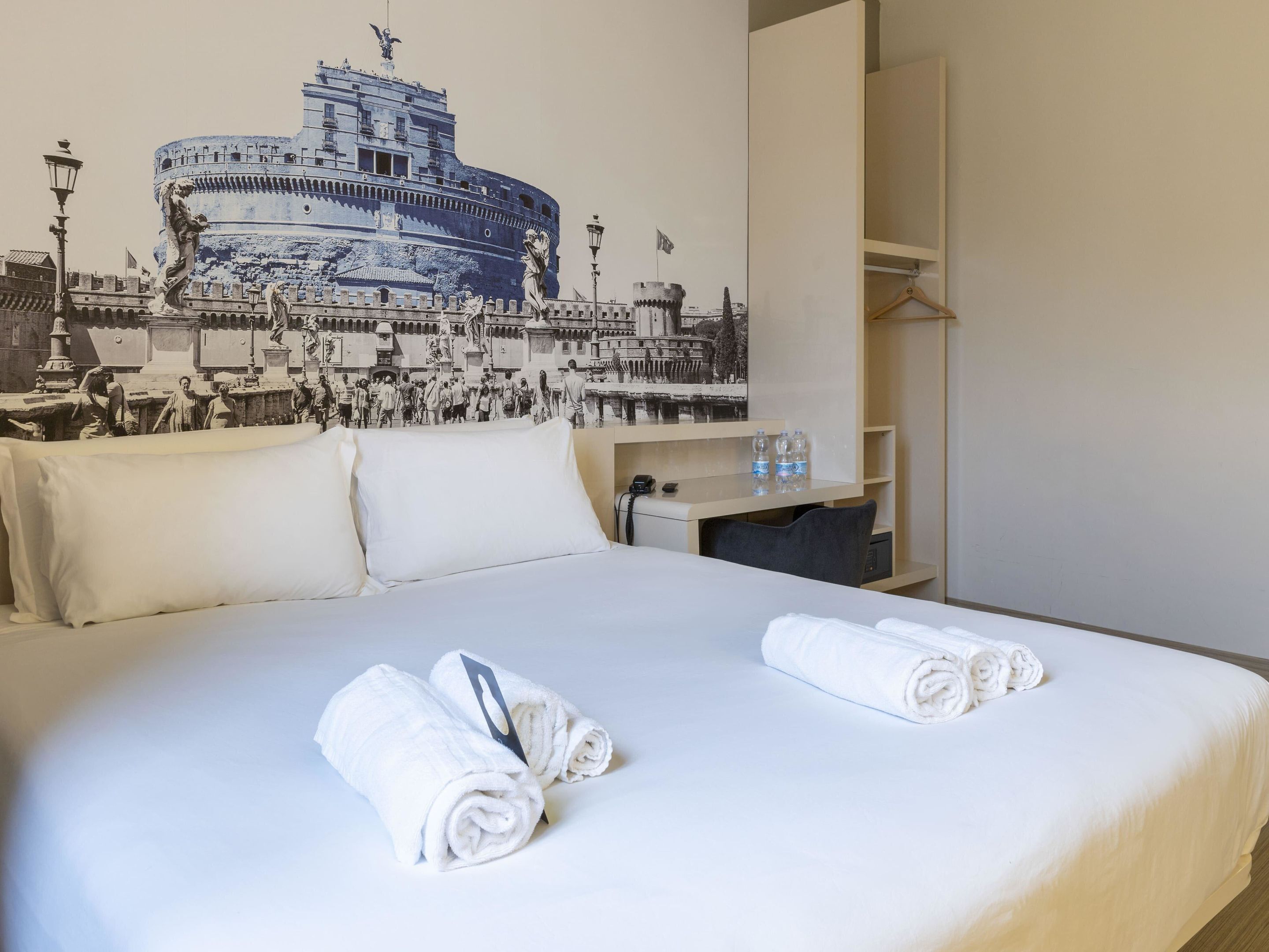 Une chambre d’hôtel avec un lit, des oreillers, une table de chevet, une bouteille d’eau et une serviette.