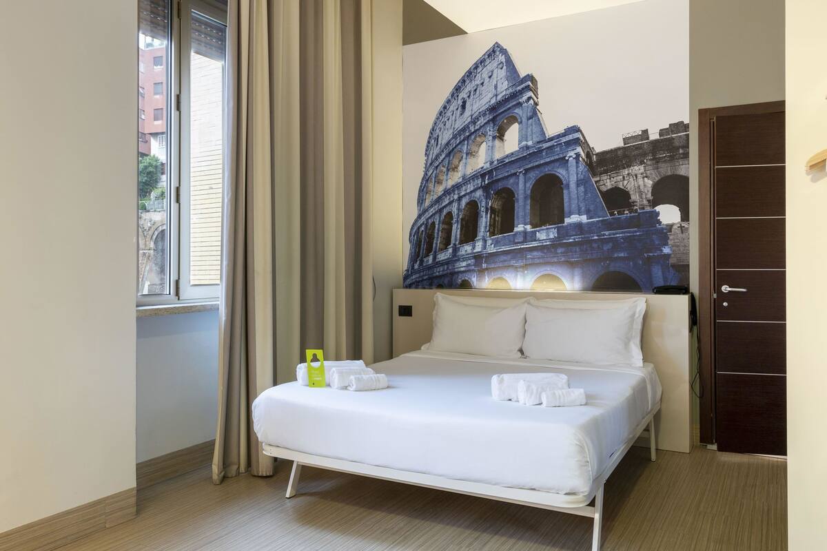 A hotel room with a bed, a window with curtains, a wall with a large architectural mural, and a door.