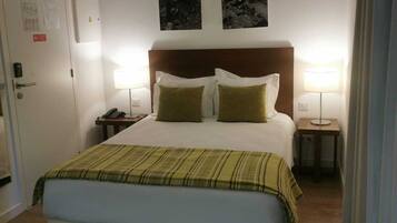 Quarto de hotel com cama, duas mesas de cabeceira, um criado-mudo, uma porta com aviso e duas quadros na parede.