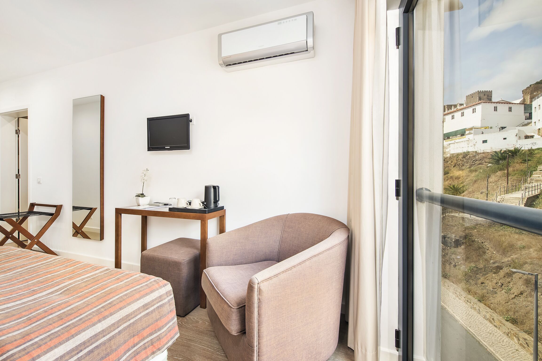 A hotel room with a bed, a chair, a small table with a kettle, a TV, and an air conditioning unit.
