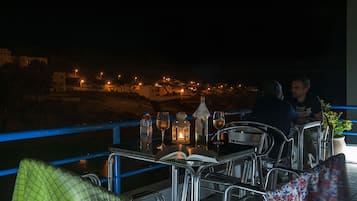 A table set with a book, glasses, and a lantern on a balcony overlooking a town at night.
