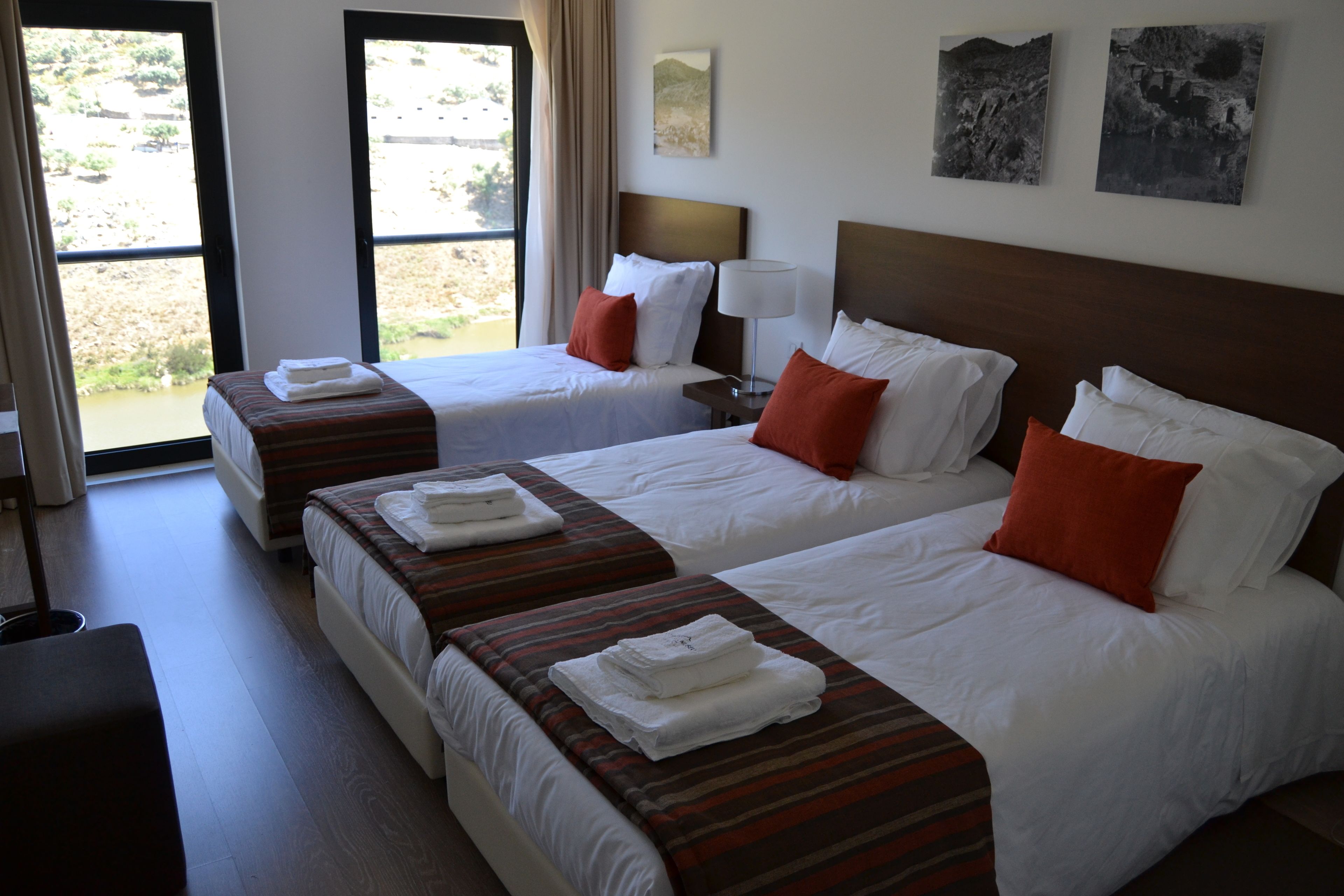 A hotel room with two beds, each with white linens and red pillows, a bedside table with a lamp, and a view of the outdoors through a large window.