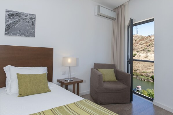 A hotel room with a bed, a chair, a nightstand with a lamp, a window with a view of a river and hills, and an air conditioning unit.
