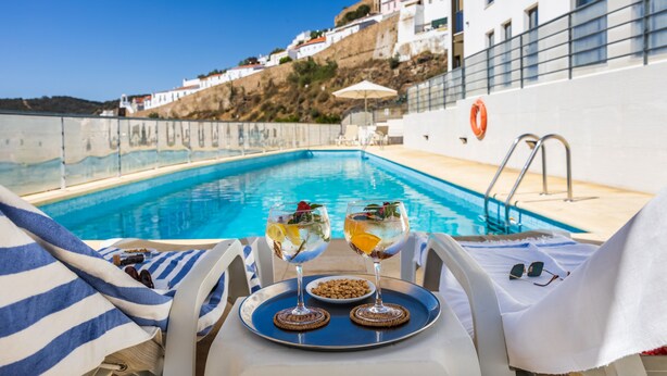 Vista de la piscina con dos vasos de bebida, una bandeja con un tazón de frutos secos y un par de gafas de sol sobre una silla.