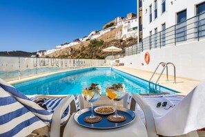 Une vue sur la piscine, avec deux verres remplis d’une boisson, un plateau contenant un bol de noix et une paire de lunettes de soleil posée sur une chaise.