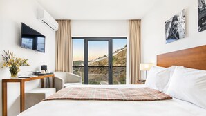 Habitación de hotel con una cama grande, un televisor montado en la pared, un escritorio con un jarrón de flores y vistas a una ladera a través de un ventanal.
