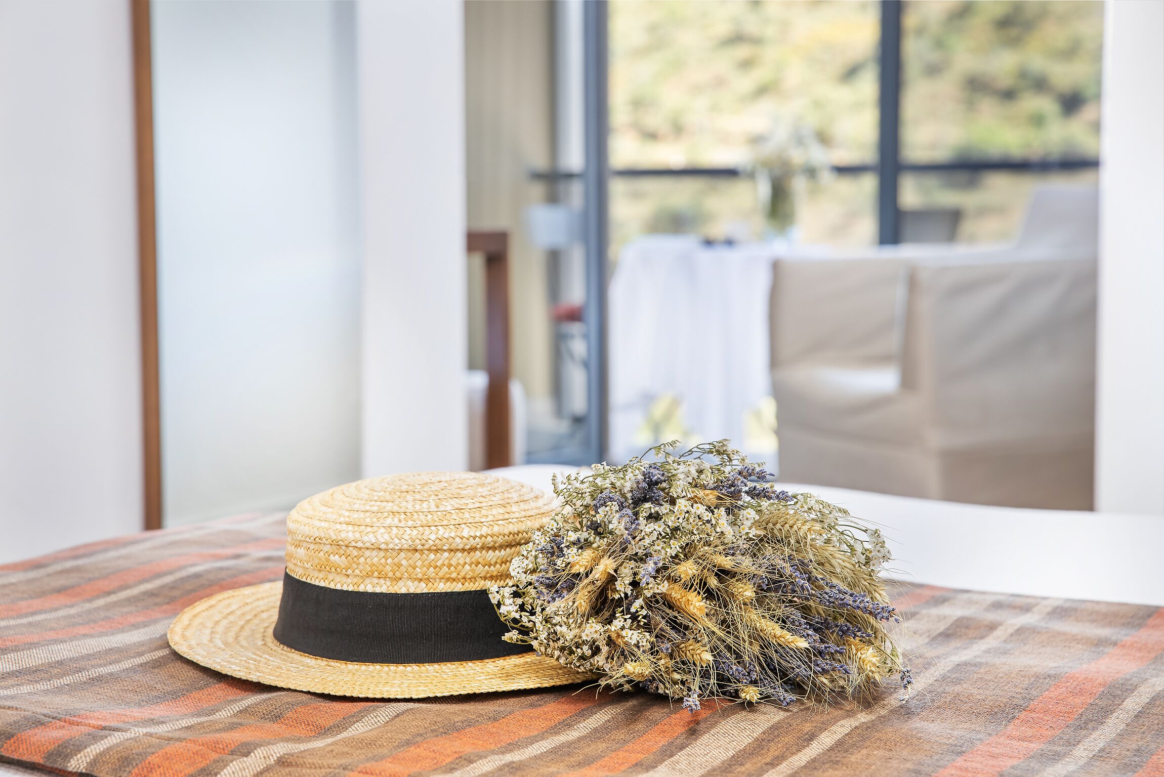A straw hat and dried flowers on a striped cushion in a room with a large window.