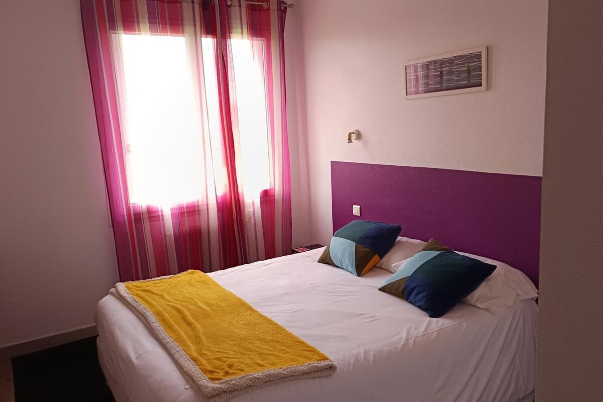 A hotel room with a bed, a bedside table, a window with curtains, and a wall-mounted light switch.