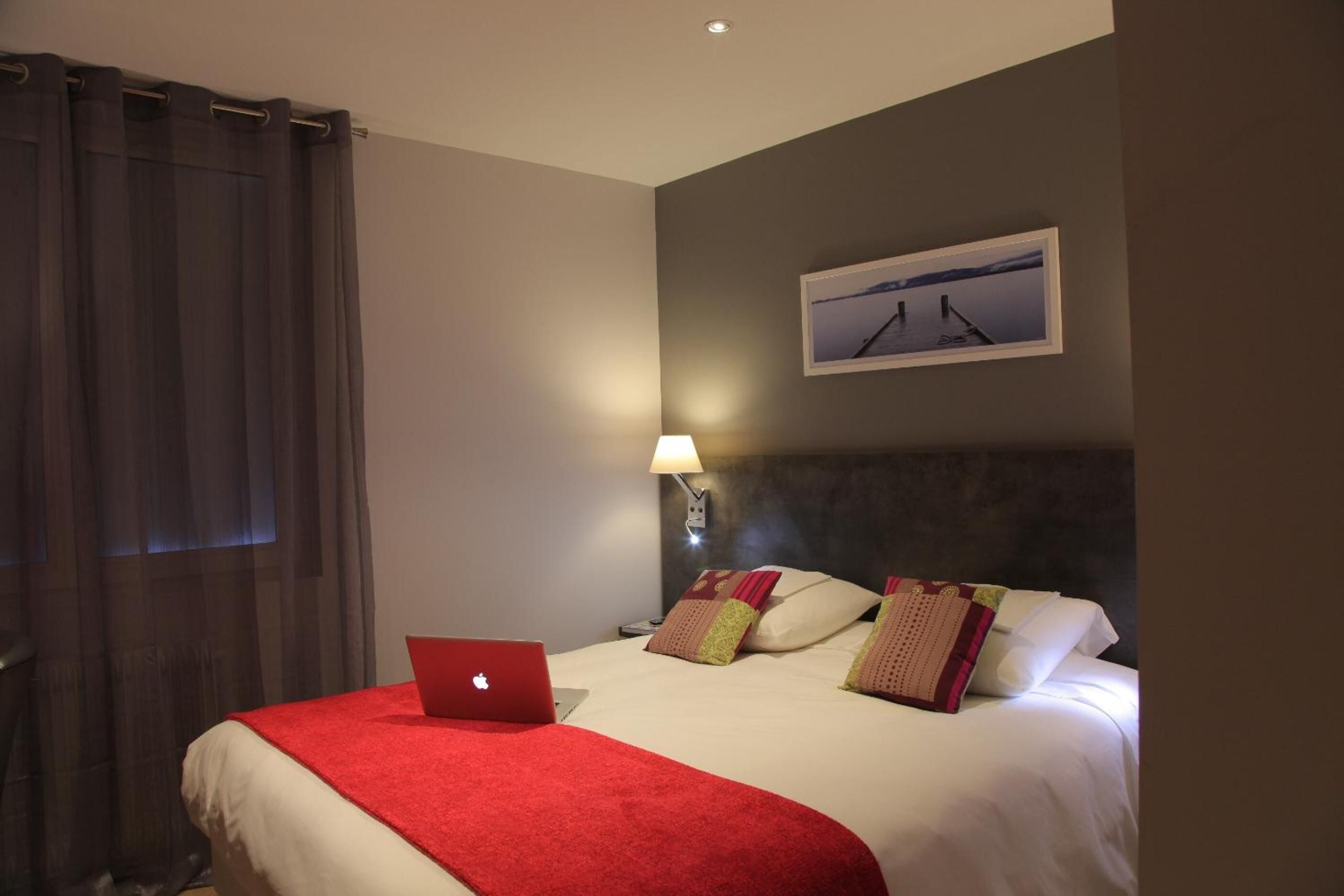 A hotel room with a bed, a laptop, a lamp, and a painting on the wall.
