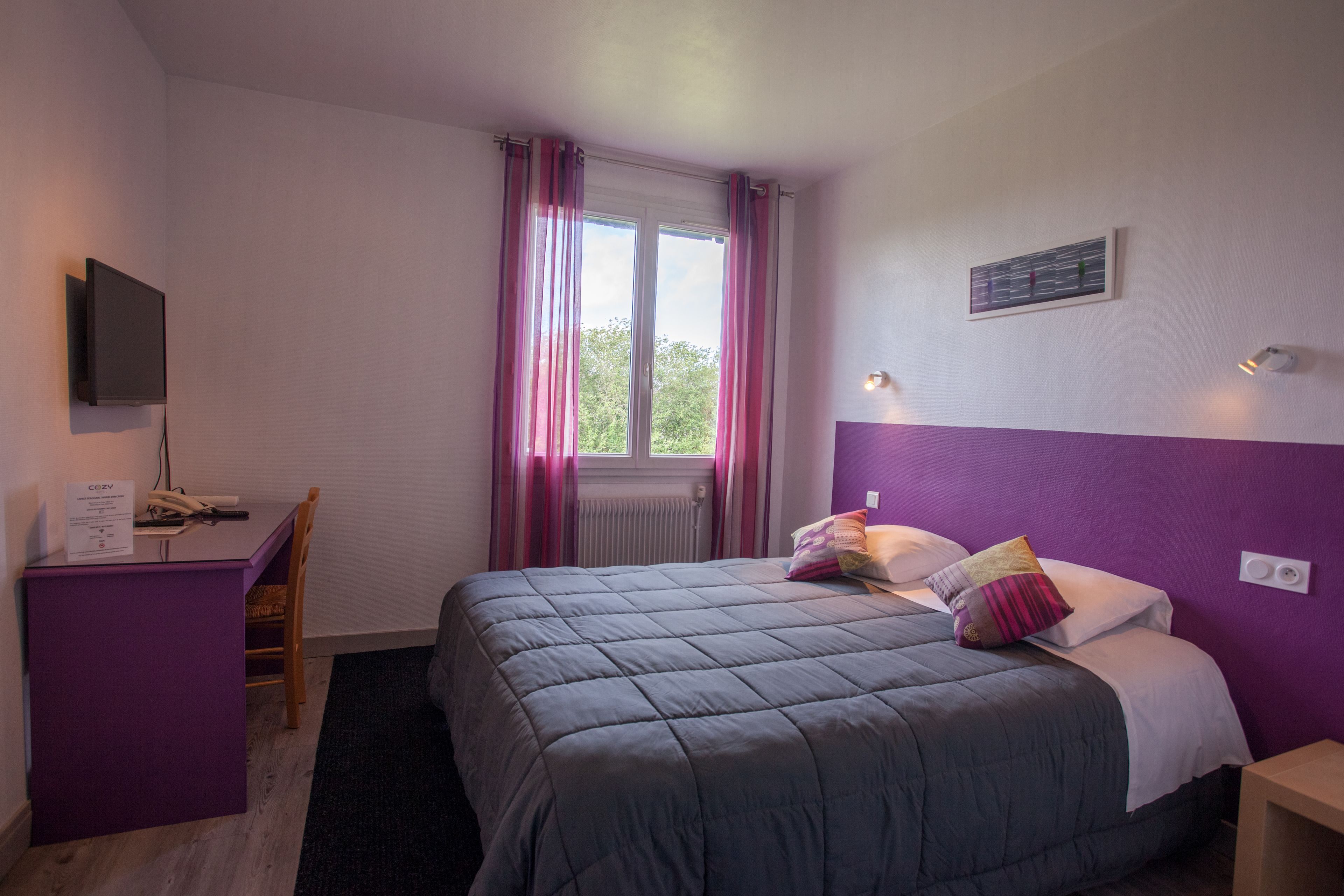 A bedroom with a bed, a desk, a chair, a TV, and a window with curtains.
