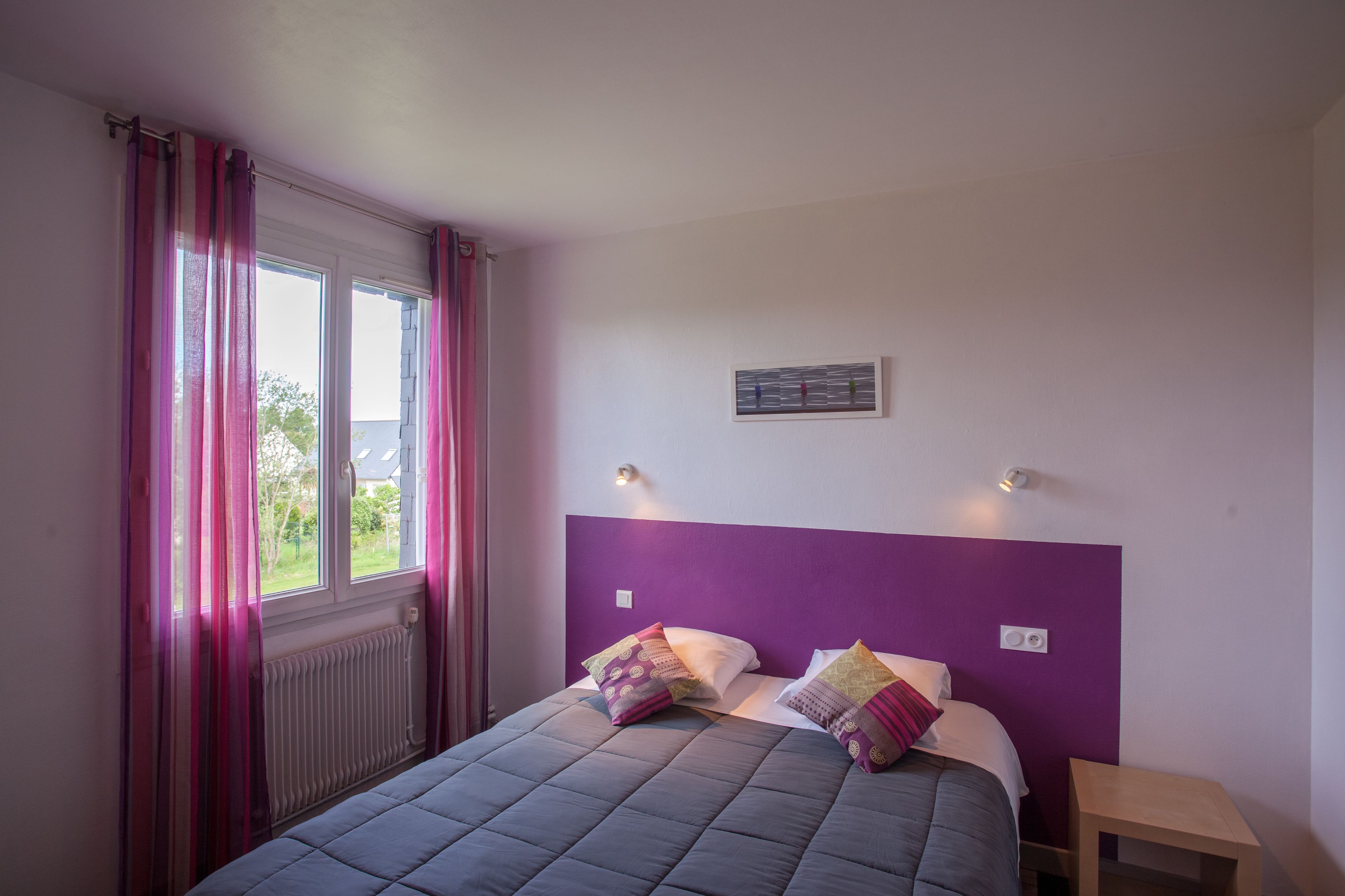 Un dormitorio con cama, cabecera morada, dos lámparas de noche y una ventana con cortinas.