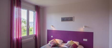 A bedroom with a bed, purple headboard, two bedside lamps, and a window with curtains.