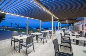 An outdoor dining area with tables and chairs, a pergola with lights, and a view of the sea. - Kymothoe Elite Suites (Zakynthos)
