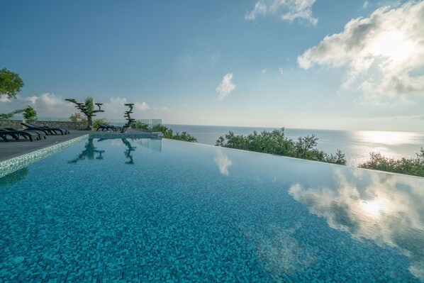A clear view of an infinity pool with a tiled edge, surrounded by lounge chairs and a view of the sea. - Kymothoe Elite Suites (Zakynthos)