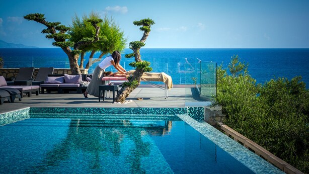 Una piscina infinita con vistas claras al mar, rodeada de sillones de descanso y un jardín cuidado.