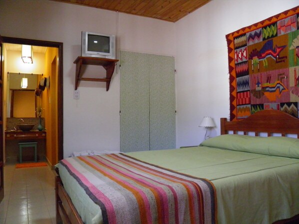 A bedroom with a bed, a television on a shelf, a colorful tapestry, and a bathroom visible through an open door.
