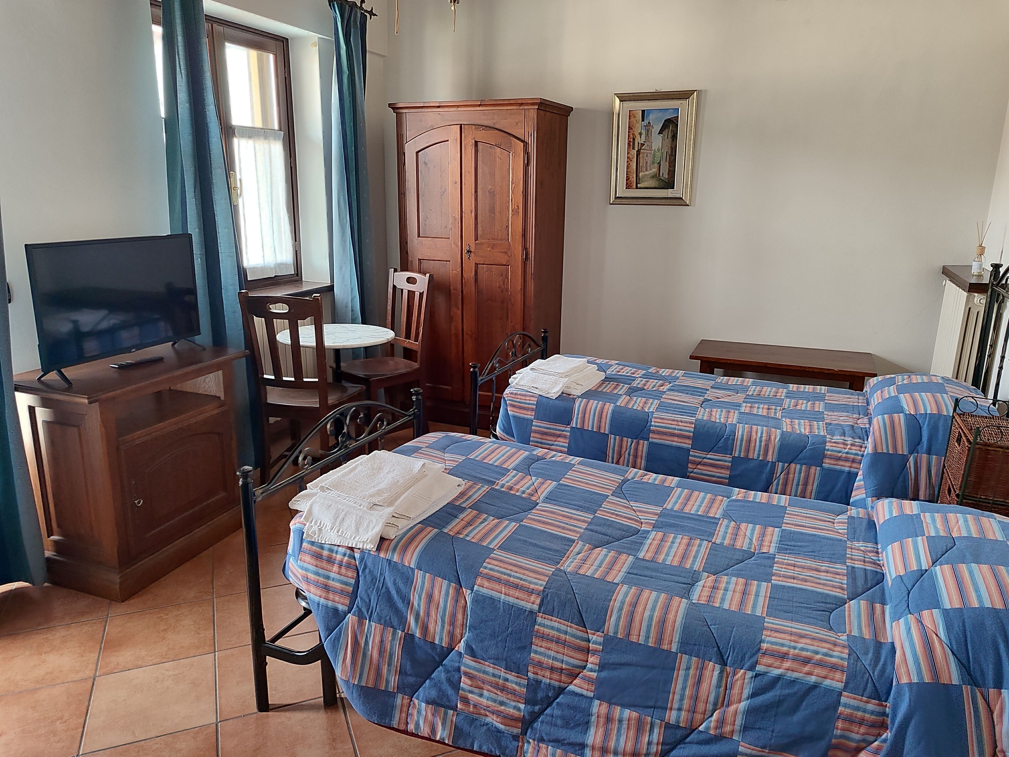 A bedroom with two beds, a wooden wardrobe, a TV on a stand, a small table, and a chair.