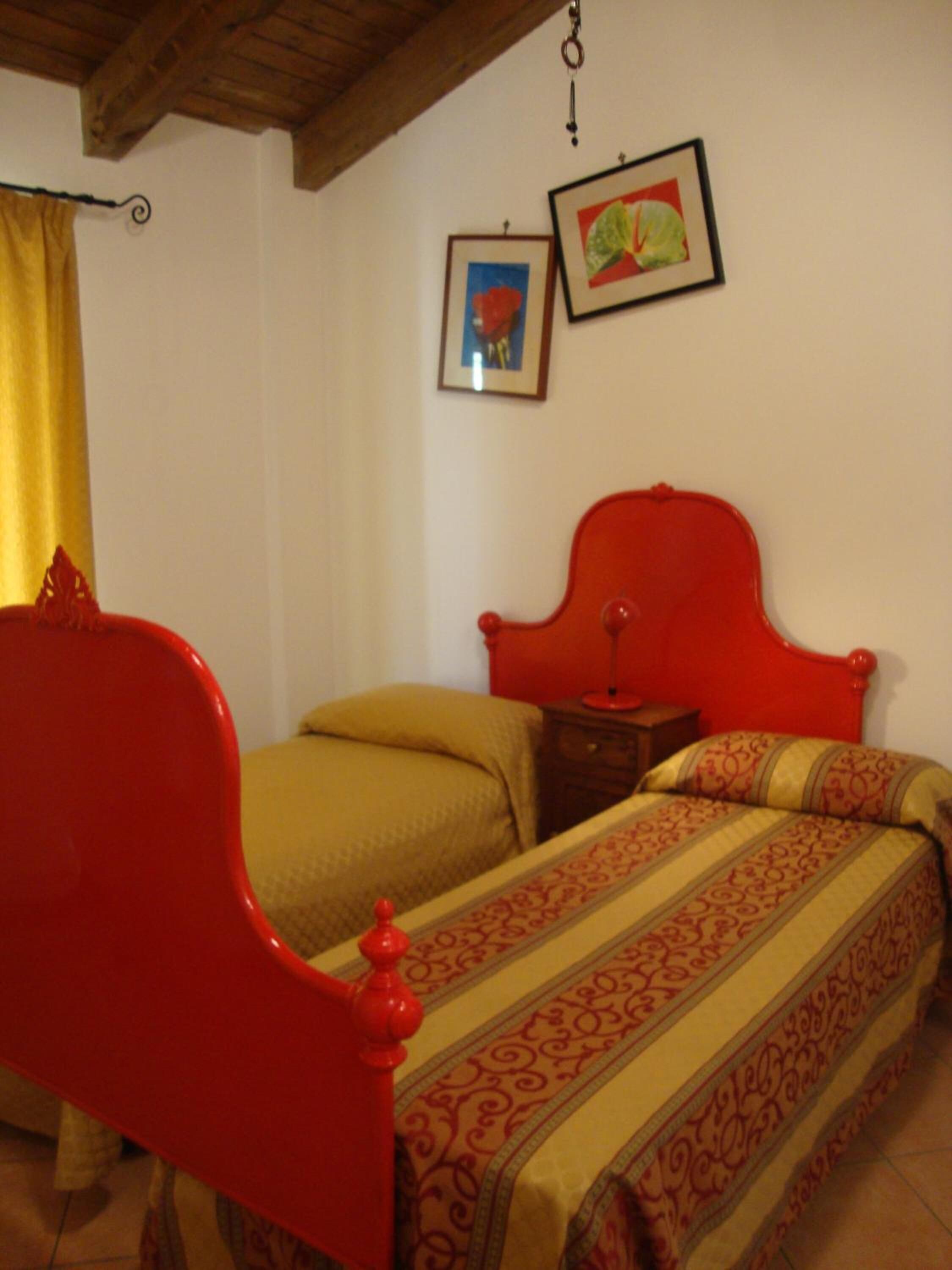 A room with two beds, one with a red headboard and another with a yellow headboard. There are two framed pictures on the wall.