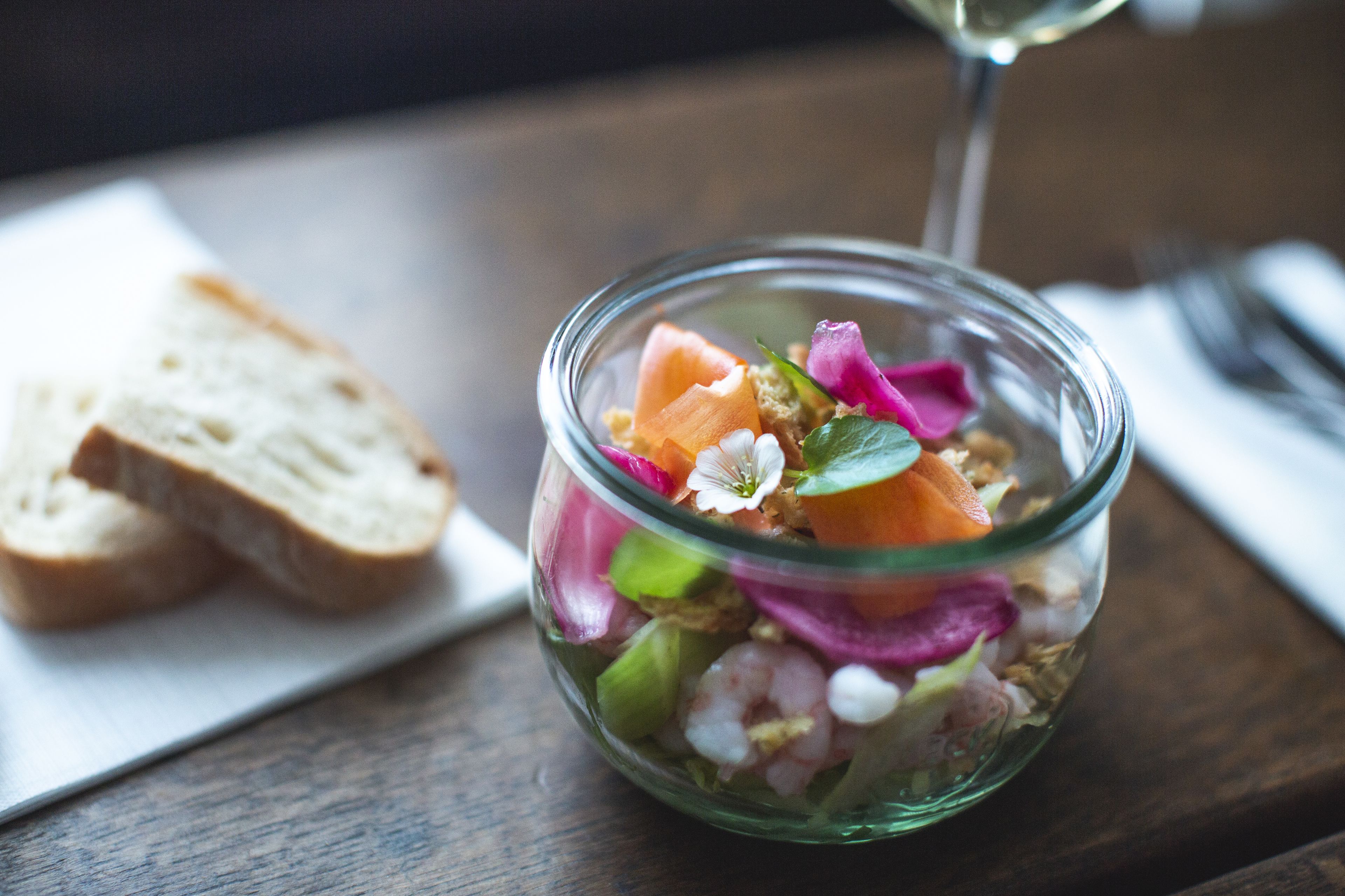 A clear glass bowl containing a salad with various ingredients, accompanied by slices of bread on a white napkin.