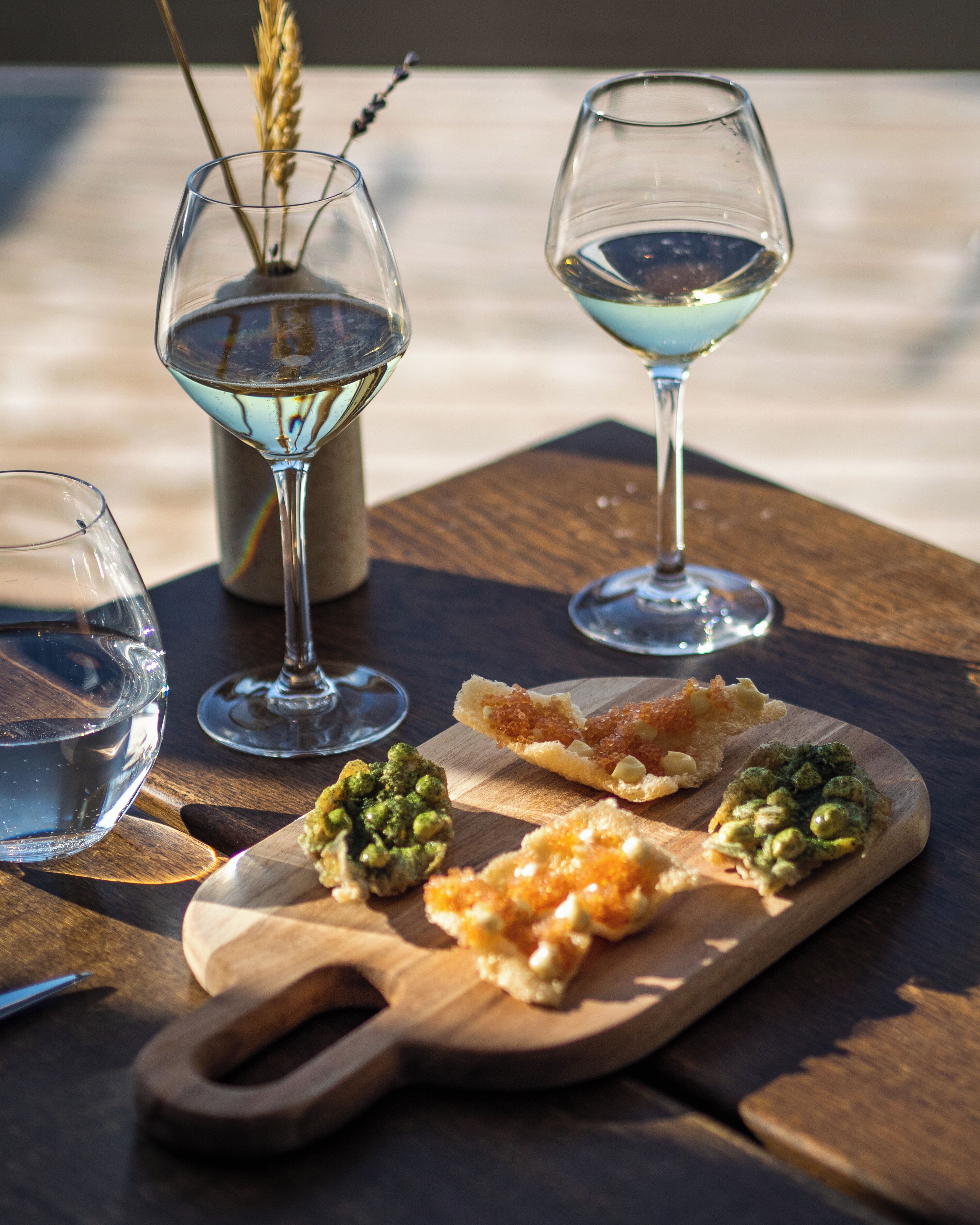 Une table en bois avec trois verres de vin, une planche en bois sur laquelle sont disposés des aliments et un vase contenant des tiges de blé.