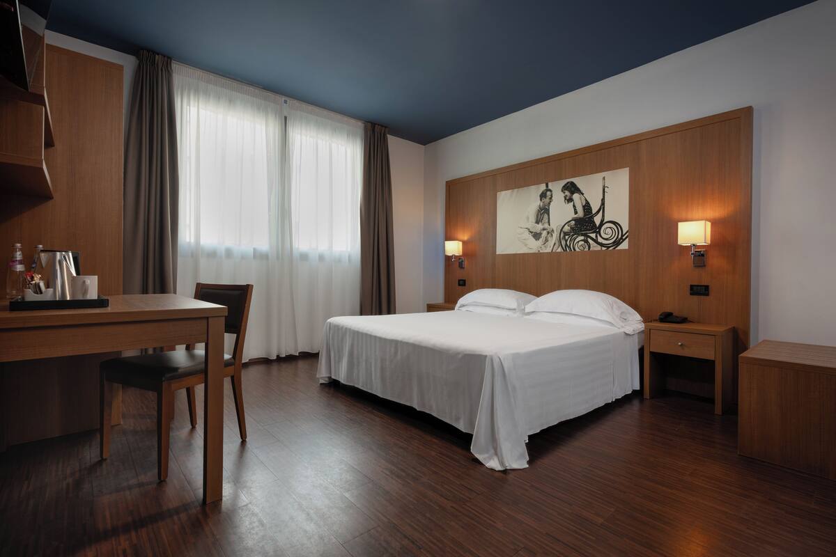 A hotel room with a bed, a desk, a chair, a nightstand, and a wall-mounted lamp.