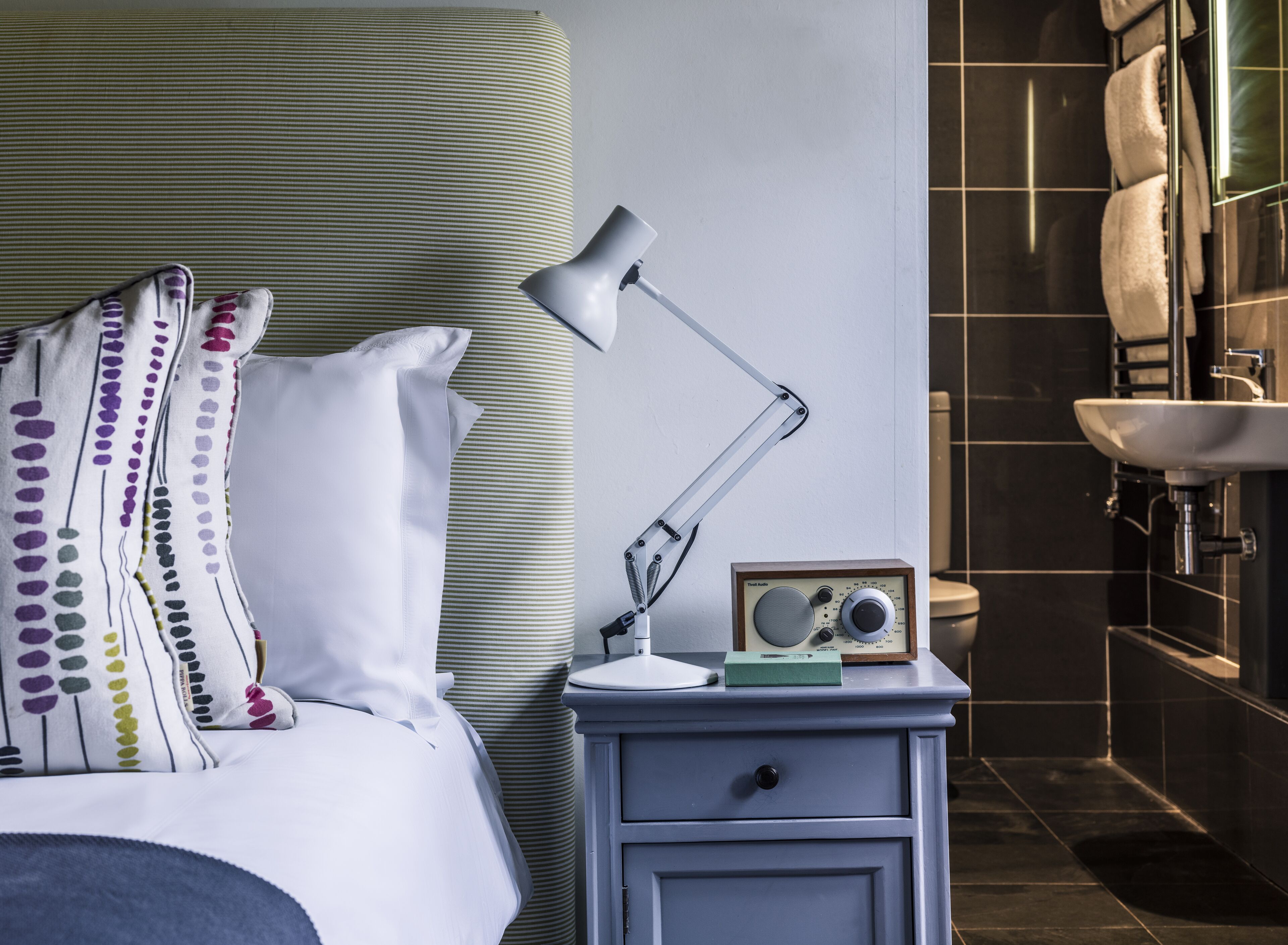 Una cama bien hecha con almohadas estampadas, una lámpara de noche y una mesita con un radio.