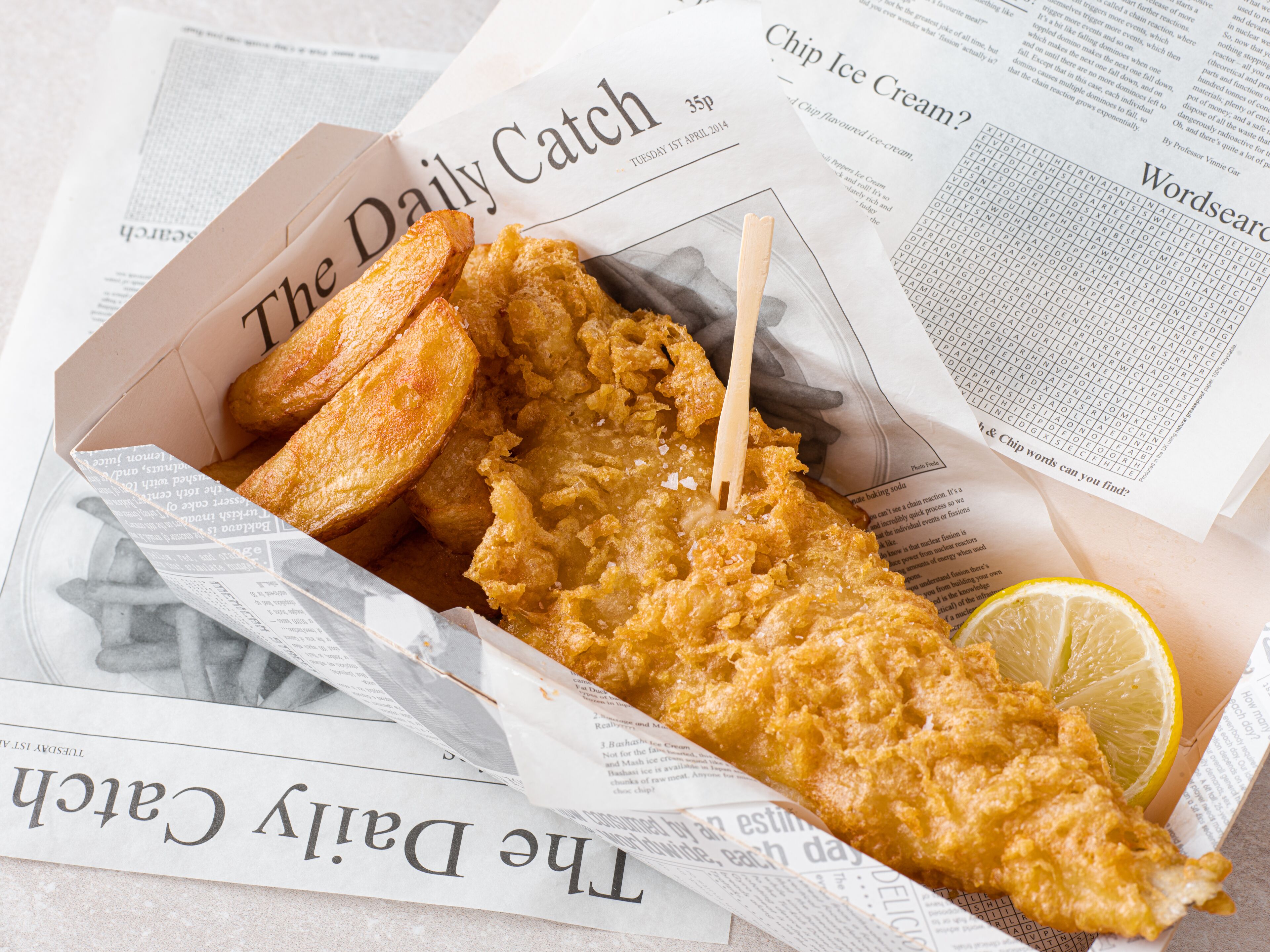 A box of fish and chips with a lemon wedge and a newspaper.