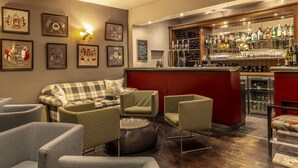 Un espace bar avec un comptoir rouge, différents types de sièges, notamment un canapé et des fauteuils à motifs, et un mur décoré de tableaux encadrés.