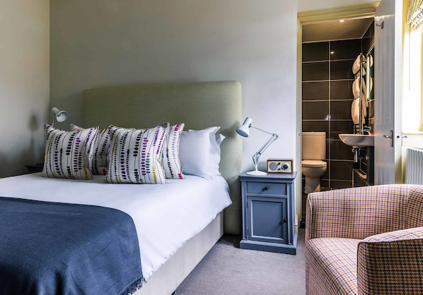 A neatly arranged hotel room with a bed, bedside table with a radio, a chair, and a bathroom visible through an open door.
