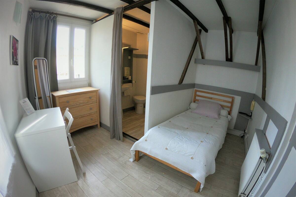 A bedroom with a bed, a desk, and a chair. There is a window with curtains, a radiator, and a door leading to a bathroom.