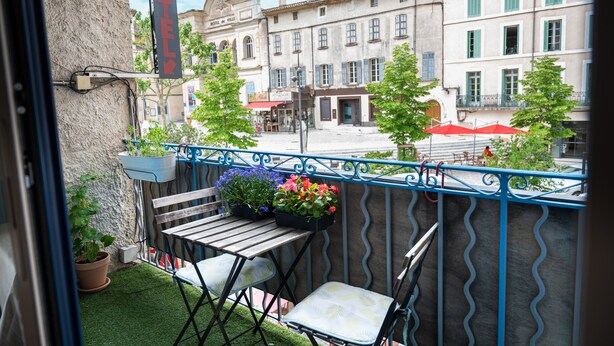 A balcony with a table and chairs, potted plants, and a view of a hotel and buildings. - Hôtel Restaurant le Palais (Apt)