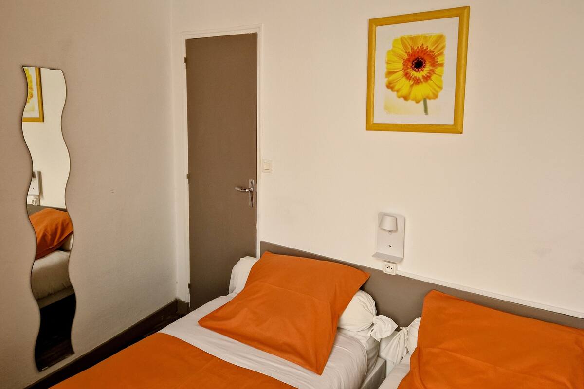 A hotel room with a bed, orange bedding, a door, a wall-mounted light switch, and a framed picture of a flower.