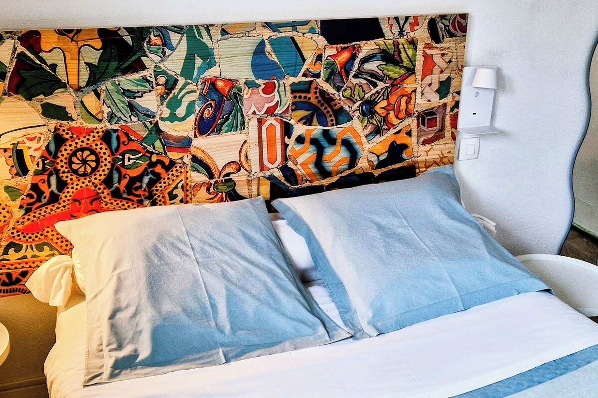 A double room with a colorful abstract headboard, a bedside table, and a chair.