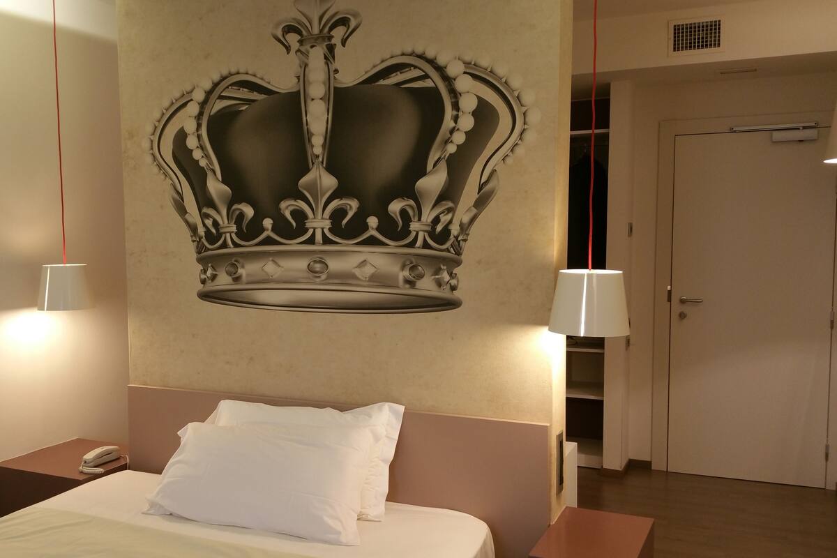 A hotel room with a large crown mural on the wall, a bed with white linens, a bedside table with a phone, and a door leading to a closet.
