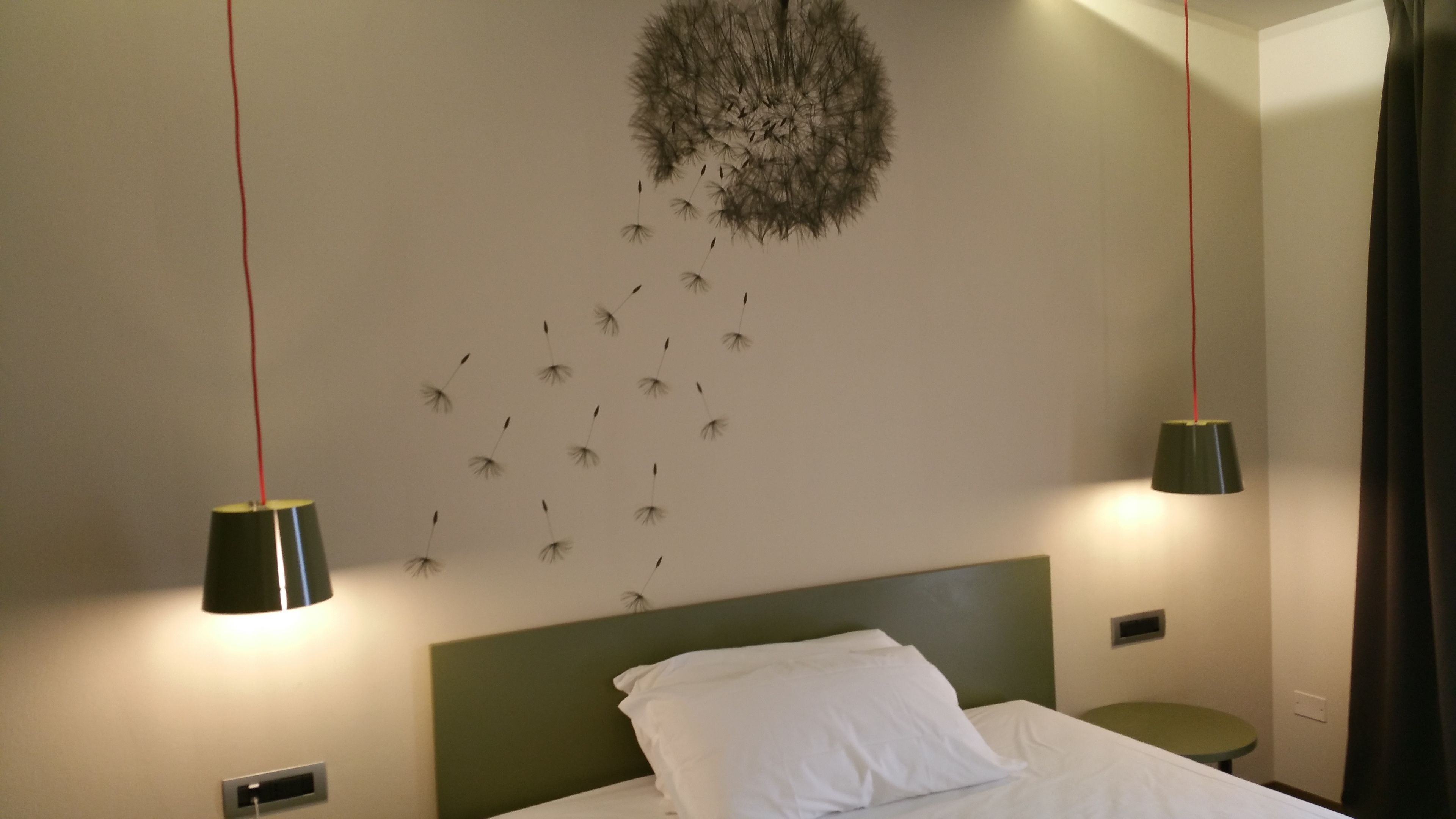 A hotel room with a bed, a green headboard, a wall art of dandelions, and two pendant lights.