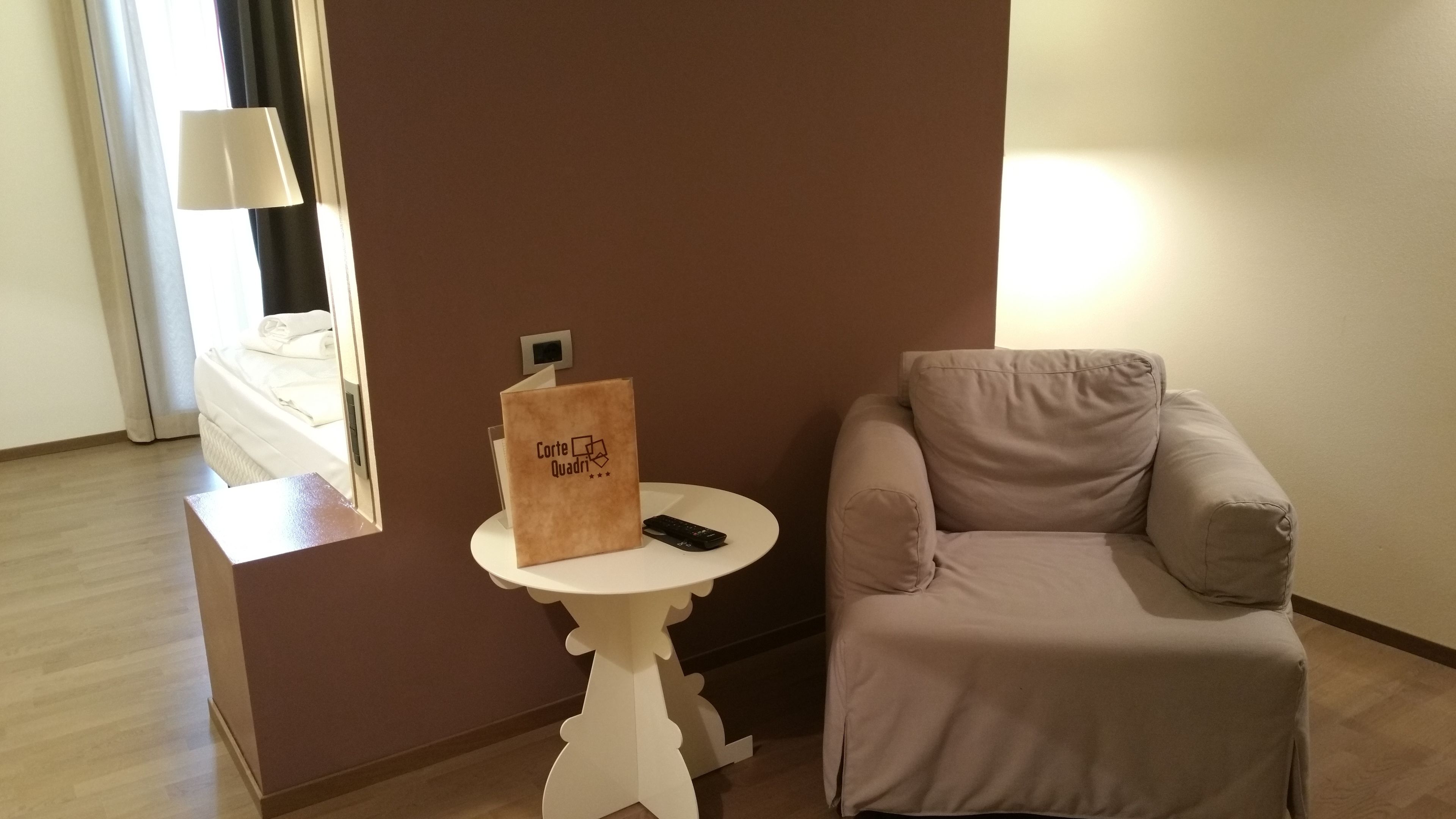 A room with a beige armchair, a small white table with a remote control and a brown paper bag, and a window with curtains.
