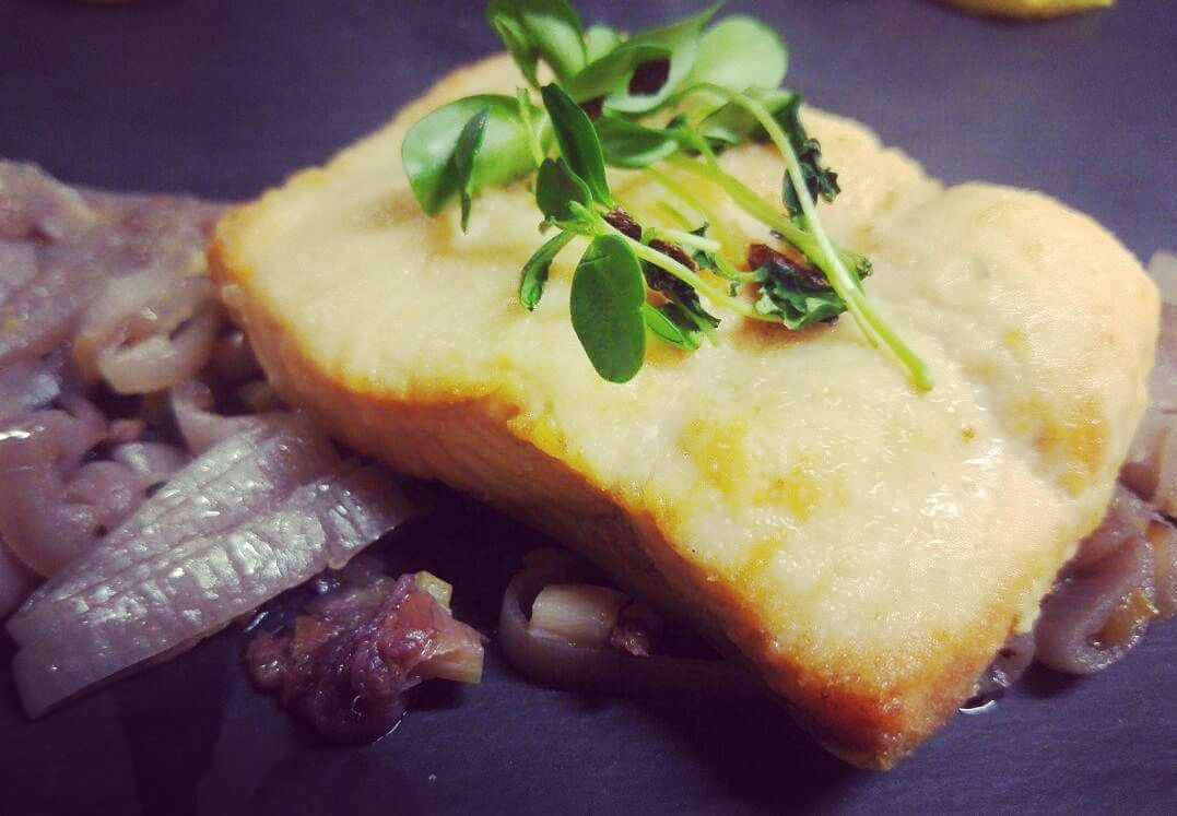 Un plato con corteza dorada, adornado con hojas verdes, servido sobre un plato oscuro junto a cebollas en rodajas y un trozo pequeño de carne.