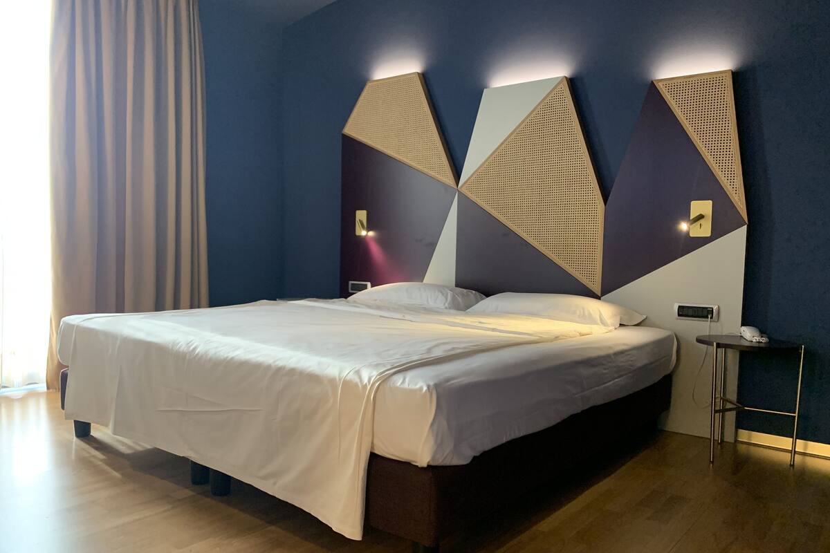 A modern hotel room with a large bed, dark blue walls, geometric wall art, and a small bedside table with a phone.
