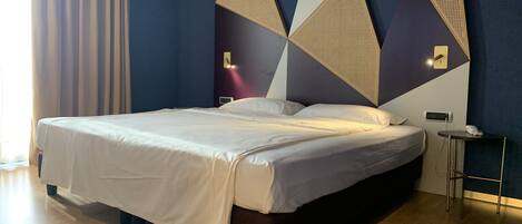 Una habitación de hotel moderna con una cama grande, paredes azul oscuro, arte mural geométrico y una mesita de noche pequeña con un teléfono.