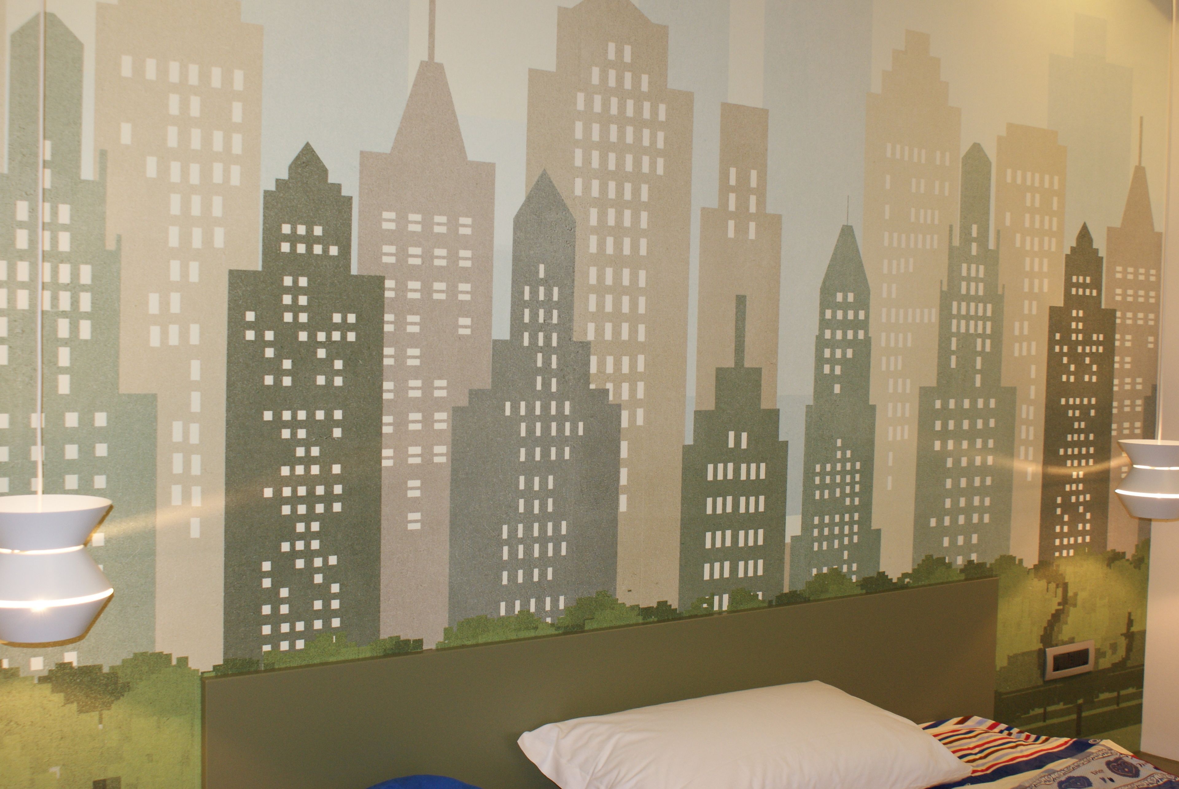 A bedroom with a cityscape mural on the wall, a bed with a white pillow, and a blue pillow.