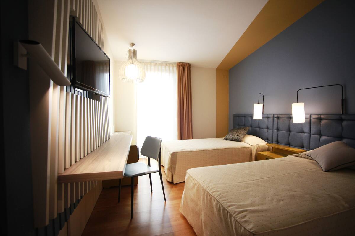 A hotel room with two beds, a desk, a chair, a TV, and a lamp.