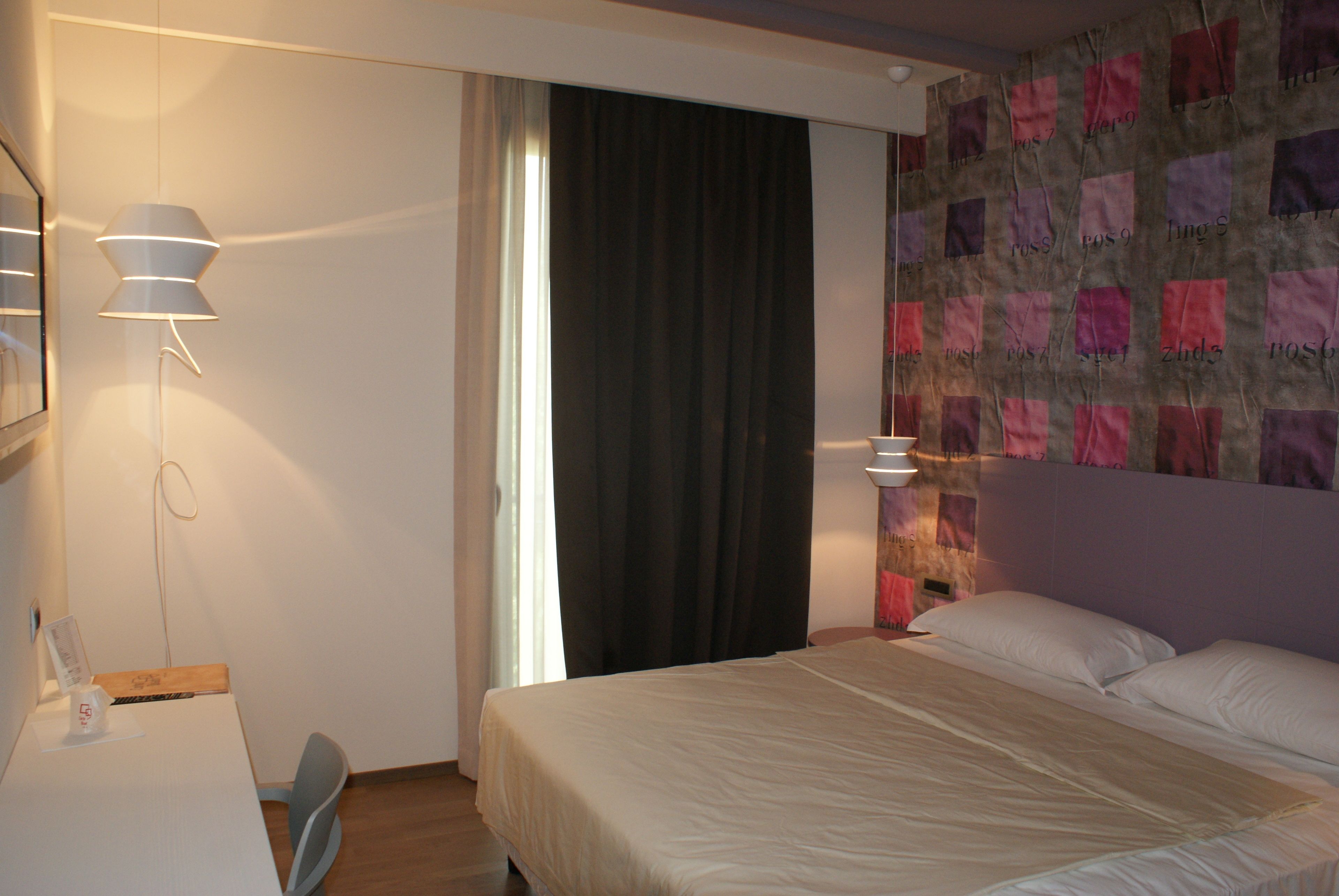 A hotel room with a bed, a desk, and a chair. There is a window with curtains, a wall with a patterned design, and a lamp on the desk.