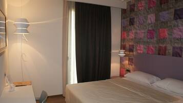 A hotel room with a bed, a desk, and a chair. There is a window with curtains, a wall with a patterned design, and a lamp on the desk.