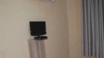 Habitación de hotel con cama, televisor de pantalla plana en la pared, unidad de aire acondicionado sobre el televisor y cortina en la ventana.