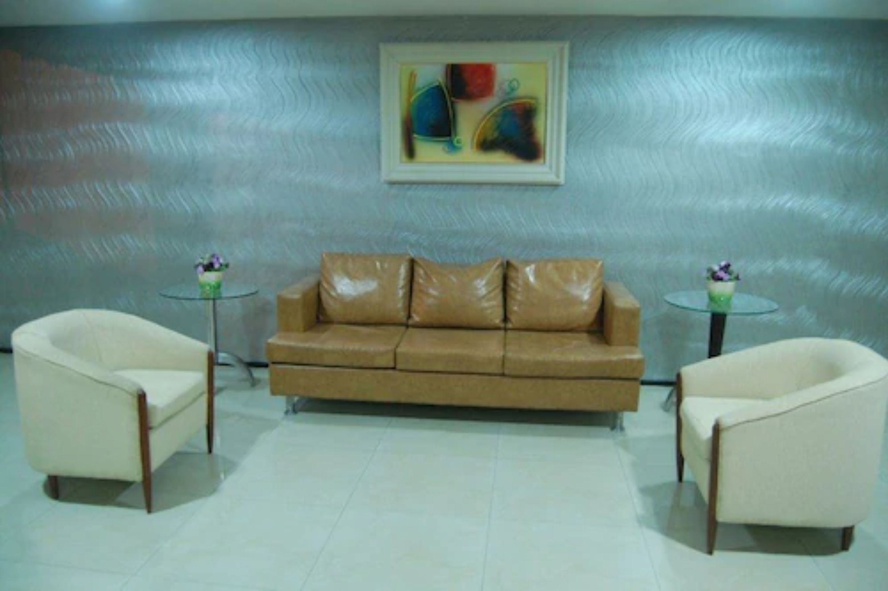 A room with a brown leather sofa, two white armchairs, a glass coffee table, and a painting on the wall.