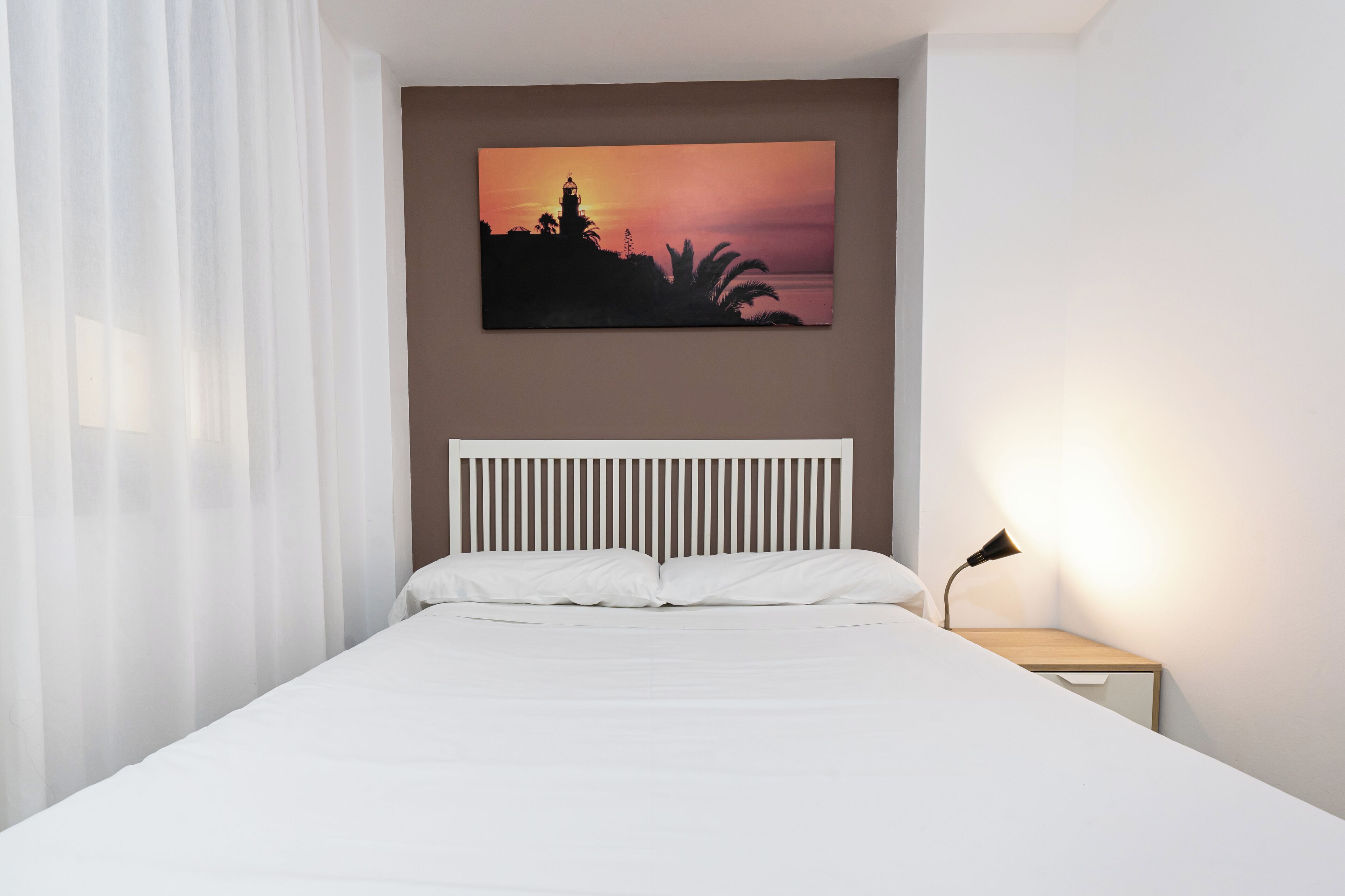 A bedroom with a bed, a nightstand, a lamp, and a sunset painting on the wall.
