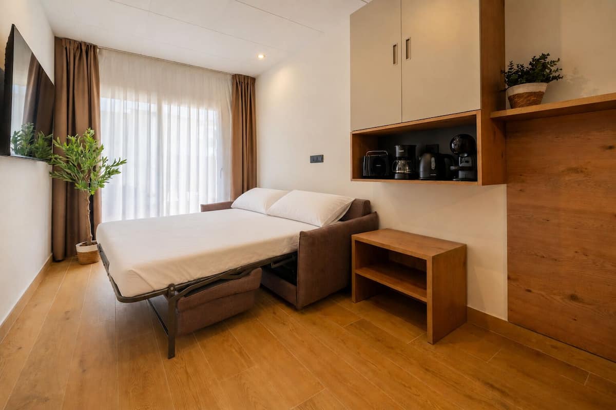 Quarto compacto com cama, uma mesinha, prateleira com cafeteiras e um vaso de planta.