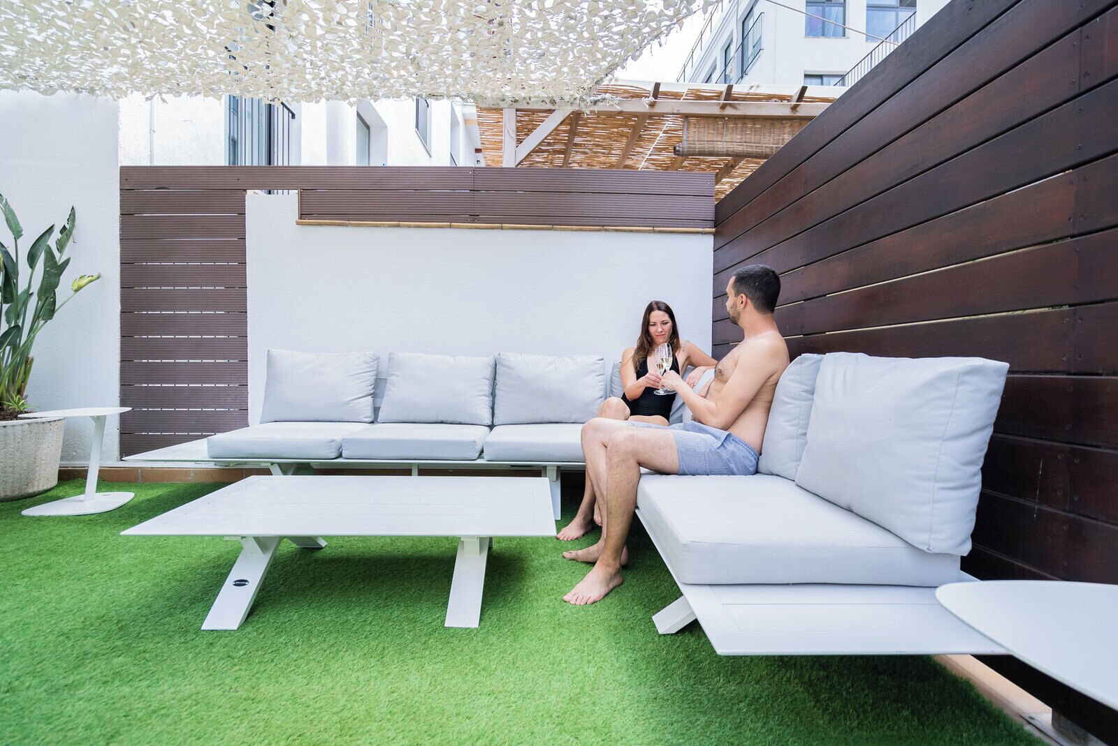 Een modern terras met een bank en twee witte tafels, omgeven door groen kunstgras en een houten wand.