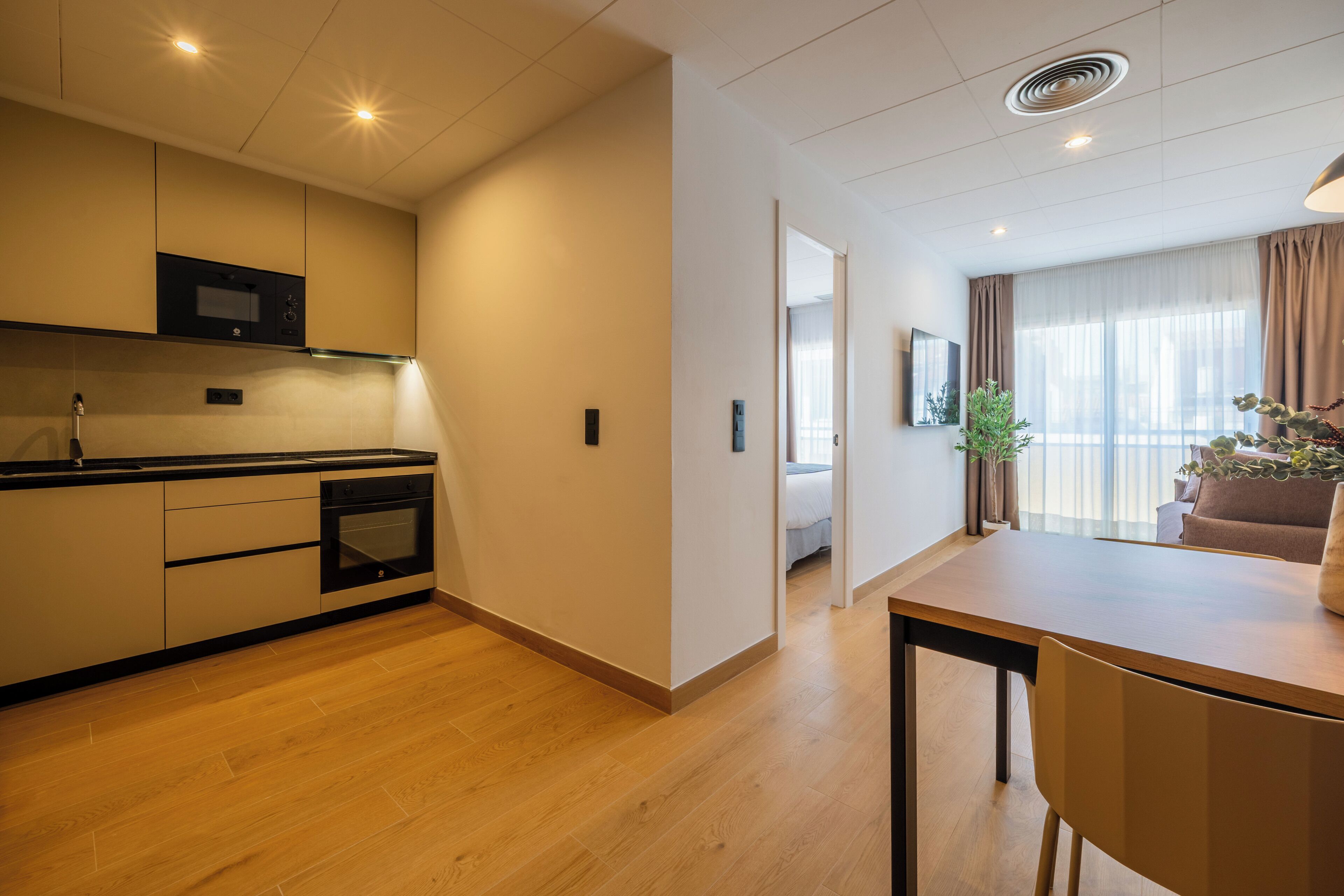 A modern kitchen with built-in appliances, a dining area with a table and chairs, and a view of a bedroom through an open doorway.